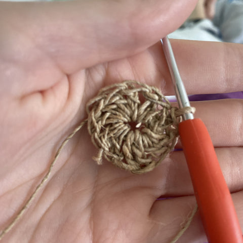 A hand holding an orange crochet hook and the beginnings of the hippie hemp coaster.