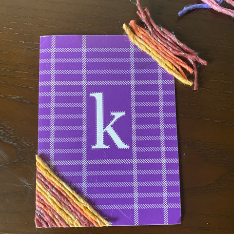 A purple plaid card with a K at the center is being upcycled with shiny orange worsted weight silk yarn