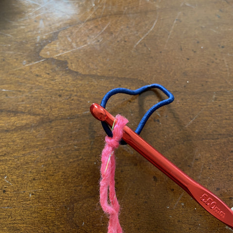 A red metal crochet hook with a navy blue hair tie has the pink sparkly yarn attached to the hook. 