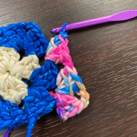 The 3rd and final yarn is being crochet onto the granny square. A white/blue/pink multicolored worsted weight yarn is being worked into the granny square's corner and side with a purple crochet hook.