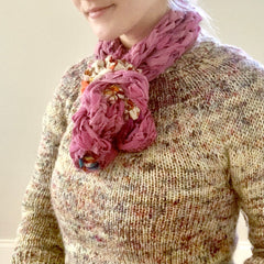 A woman is smiling, wearing a soft woven sweater and a pastel pink finger knitted scarf around her neck.