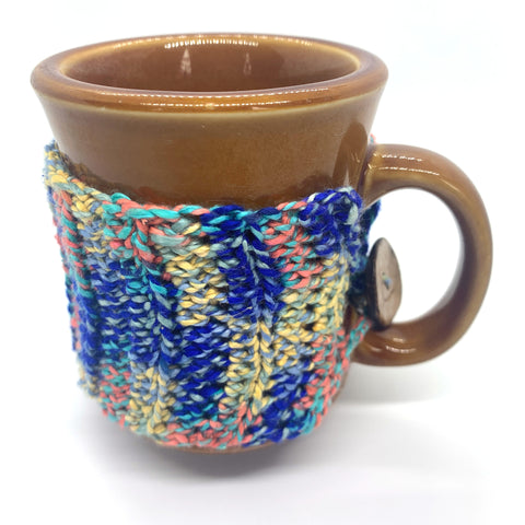 Brown coffee mug wrapped with crocheted blue yellow and peach rain boots yarn mug snug on white background. 