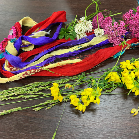 On a dark wooden desk a pile of different colored ribbon and flowers are laid out, ready to be braided and woven into a witches' ladder.