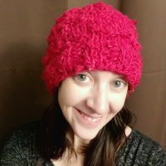 A pale woman with dark hair is smiling at the camera, wearing a bright fushia knit beanie.