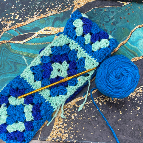 A headband made of blue granny squares is laying on a green, black, and gold marble cover. To the right is a ball of teal worsted weight yarn and a golden crochet hook.