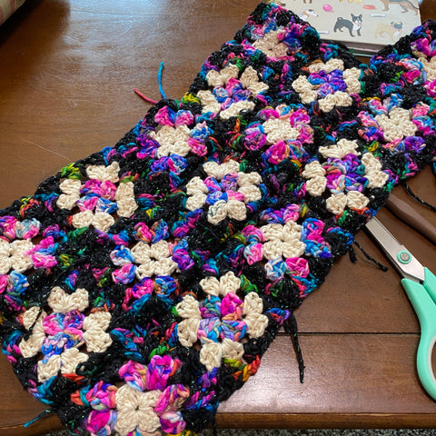 Three lines of multicolored granny squares are stitched together and laying on a wooden table by a pair of seafoam green scissors.