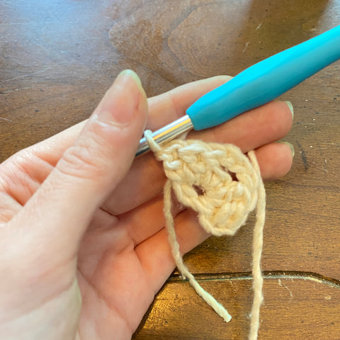 a pale hand is holding a half finished center of a granny square, made of white worsted weight yarn. A light blue crochet hook is still attached to the yarn.