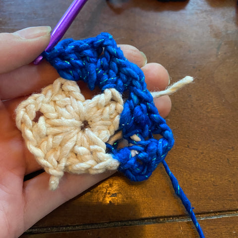 A pale hand is holding a half finished granny square, the two right corners of the granny square filled in with sparkle classic blue worsted weight yarn.