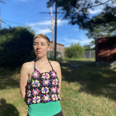 A person with short blonde hair is standing outside in the sun wearing the granny square top and a bright green tank top.