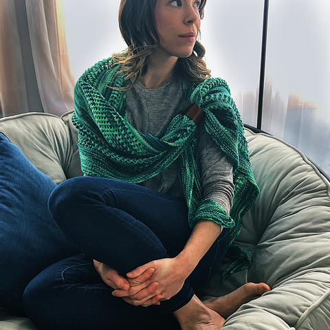 A woman with long brown hair wearing an ombre green shawl, blue jeans, and a grey shirt is curled up on a large, cushy bean bag poof.