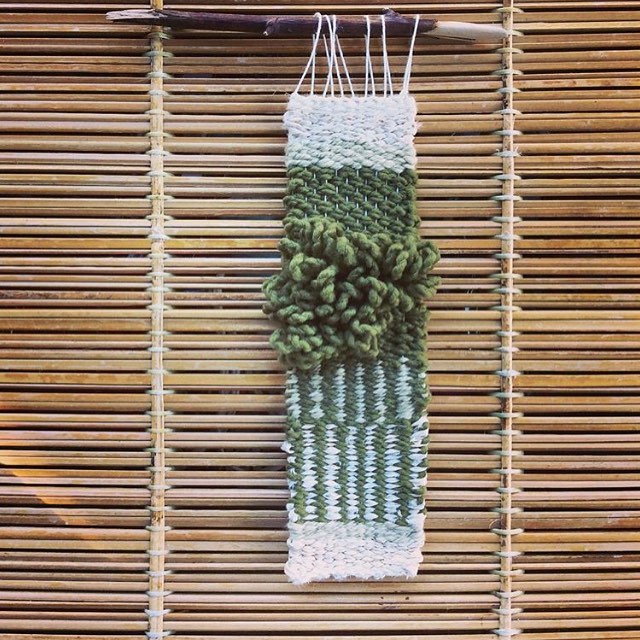 Green and white woven wall hanging hung from a branch on a wooden wall