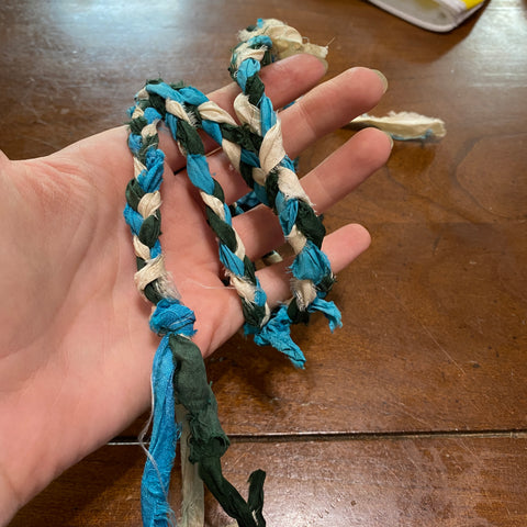 A pale hand is holding a finished handfasting cord, plaited together with white, blue, and dark green sari recycled ribbon yarn. The single plait is twisted around the person's hand.