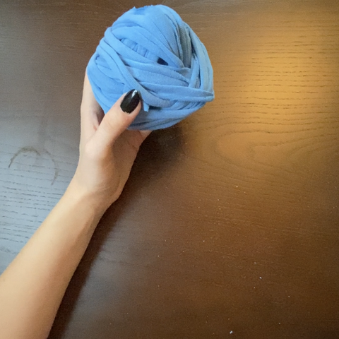 A hand with black nails is holding up a wound up ball of blue t-shirt yarn against a dark wooden table.