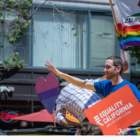 A man wearing a blue shirt holding an orange 'equality for california' sign at a priderally is waving as he marches down the street, surrounded by signs and flags.