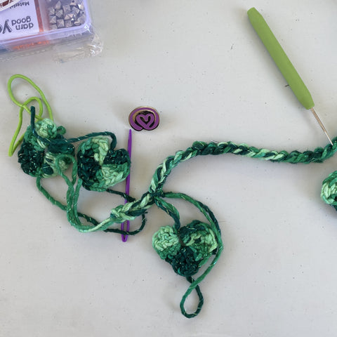A green heart garland is laying out on a white plastic table. A purple darning needle is halfway through weaving a heart into the garland chain. To the right of the image is a green loom pick and to the left of the garland is a plastic bead kit. At the point of the darning needle is a purple DGY enamel pin.