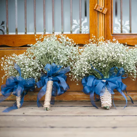 On the floor, resting up against a wooden wall, are three different bouquets of small white flowers, their stems wrapped in blue ribbons, pearls, and hemp yarn.