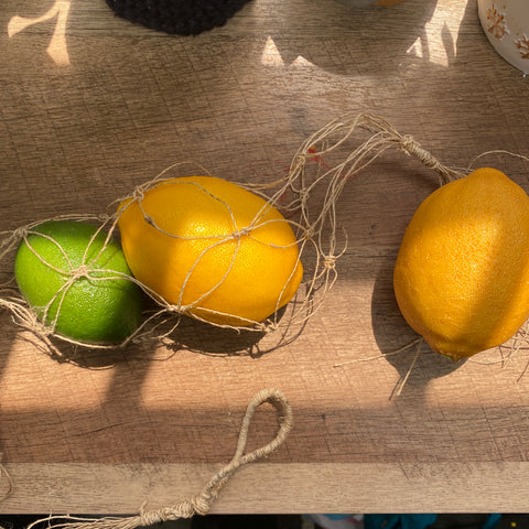 A lime and two lemons are securely held in a hemp macrame hanging produce bag as they lay on a wooden table in the sun.