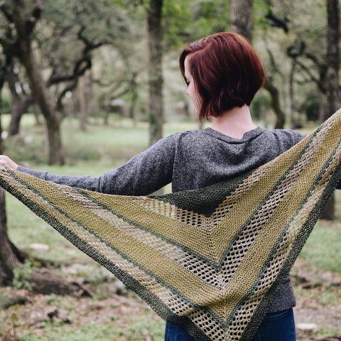A woman is in the woods, facing away from the camera, spreading her arms out to show off her ombre green knitted shawl made from DK weight silk yarn.