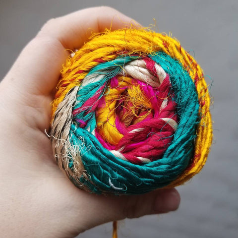 A hand wrapped around a cake of spice market yarn, made of pink, yellow, white, and teal yarn.