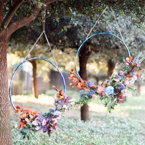 From a tree, two large blue hula hoop flower wreaths are hanging from the branches. The hoops are decorated with orange, purple, and green flowers.