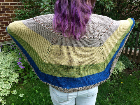 A woman with long purple hair is facing away from the camera, spreading out her tan, green, and blue knitted shawl.