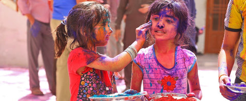 Two young girls are covered in colored powder. The girl on the left is smearing purple pigment onto the other girl's cheek