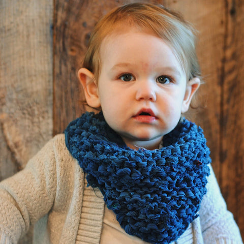 A baby is sitting, staring at the camera, a blue bandana wrapped around their neck.