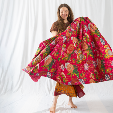 A woman with red hair and wearing a black shirt is holding up a pink kantha quilt.