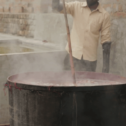 A gif of an Indian man wearing a white shirt, a black mask, black gloves, and goggles is stirring a large boiling dye pot with a long stick.