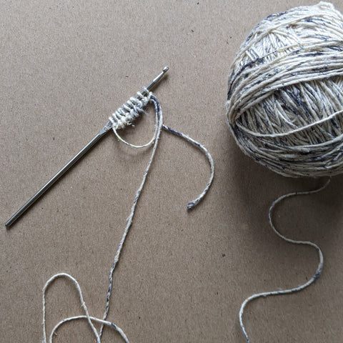 A ball of gray and white speckled yarn is attached to a silver crochet hook on a wooden table.