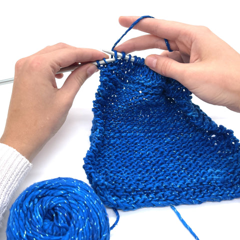 A pair of hands are working on a white table, a pair of knitting needles in their hands as they work up some sparkly blue worsted weight silk yarn.