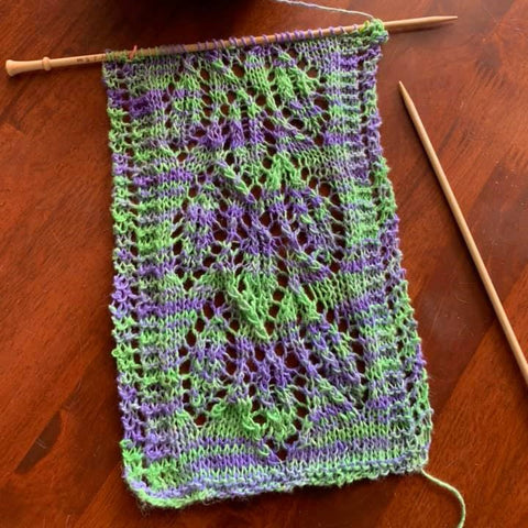 A gorgeous and intricate knit stitch pattern, made out of purple and green lace weight silk yarn is laid out on a wooden table.