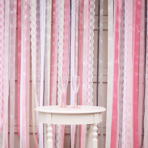 A ribbon and lace photo backdrop, made of white and different shades of pink ribbon and lace. At the center is a white table with two champagne flutes on the tabletop.
