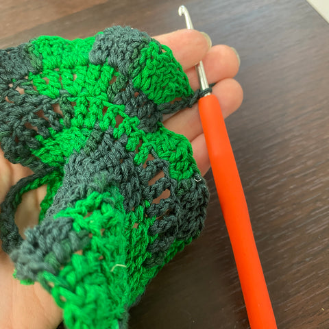 A pale hand is holding an unfinished crochet green leaf and an orange crochet hook.
