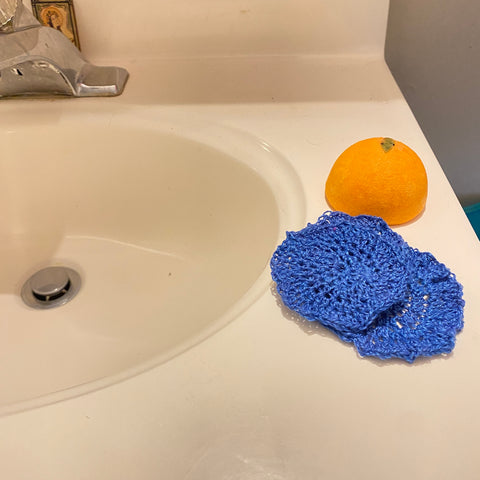 On the edge of a porcelain sink sits a pair of blue crochet makeup remover pads as well as an orange scrubbie. 