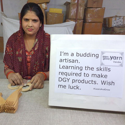 One of our artisans, Mamta, wearing a burgundy sari is sitting at a work table. To the right there is a sign that reads 'I'm a budding artisan. Learning the skills required to make DGY products. Wish me luck!'