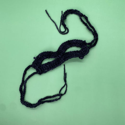 A mask made of black worsted weight silk yarn is laying on a light green background.