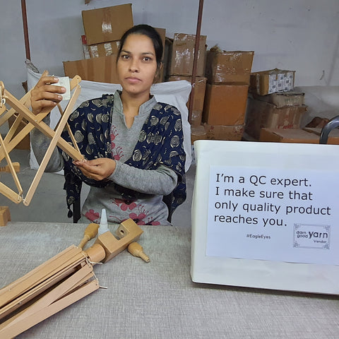 Soniya, a young Indian woman wearing gray and navy blue is sitting at a gray table. In her hands is a wooden yarn swift. To the right of the table is a white sign that reads 'I'm a QC expert. I make sure that only quality product reaches you!