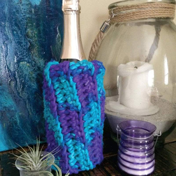 Mermaid Party Wine Sleeve on a wine bottle next to plant and candle on a wooden table in front of a blue wall