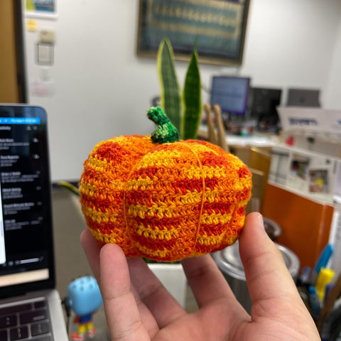 A hand is outstretched, showing off an ombre crochet pumpkin amigurumi made out of ombre orange and green yarn