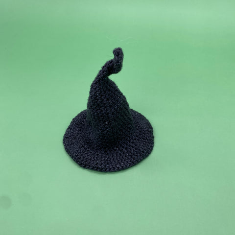 On a green background sits a little crochet witch hat made from lace weight silk yarn in black