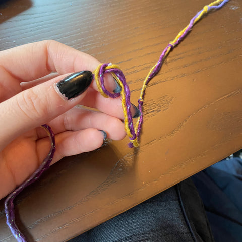 A hand with black painted nails is knotting the sparkling purple and yellow yarn