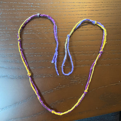 A heart made out of knotted purple and yellow sparkle yarn is forming a hart on a dark wooden table.