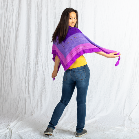 A woman with long dark hair is turned away from the camera, but is twisting her shoulders and head around to smile at the camera. She's wearing jeans, a yellow shirt, and an ombre purple triangular shawl.