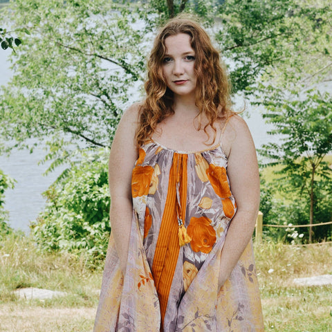 A woman with curly red hair is wearing a tan and orange maxi dress.