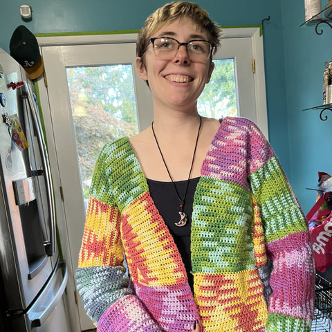 A person with short hair and glasses is wearing the multicolored worsted weight patchwork cardigan.