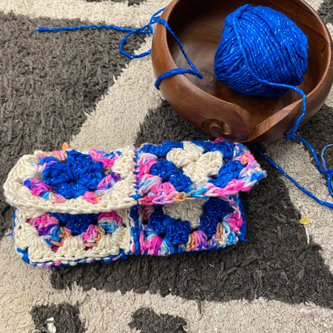 The finished multicolored granny square pouch is resting on a fluffy gray and white rug. To the right of the case is a wooden yarn bowl and a cake of sparkle classic blue worsted weight yarn.