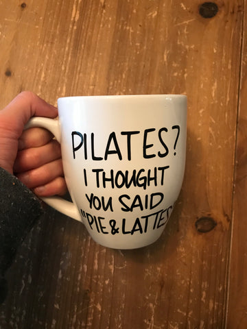 Woman holding a white mug with black text that reads 'Pilates? I thought you said Pie & Lattes' over a wooden surface