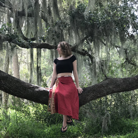 A person with short blonde hair, a black top, and a red wrap skirt, is sitting on a large tree limb in the woods.
