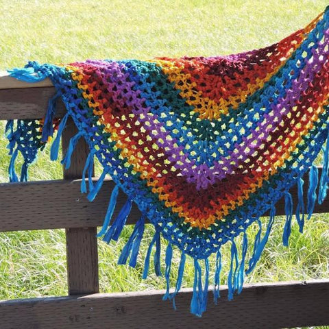 Rainbow crochet shawl draped over a wooden fence in front of a grassy area